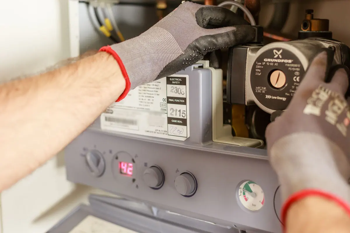 Gloved hands working on a gas boiler's circulating pump, with the boiler's controls and pressure gauge below